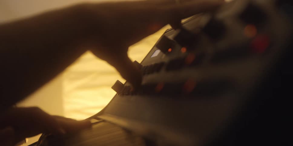 The video is a dimly lit closeup shot of hands interacting with a music synthesizer or keyboardthe hands are seen adjusting the knobs and touching the keys of the instrument which has several illuminated orange buttons and controls
