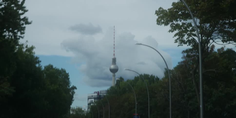 A famous television tower rises above a tree lined street in a cityscape under a cloudy sky