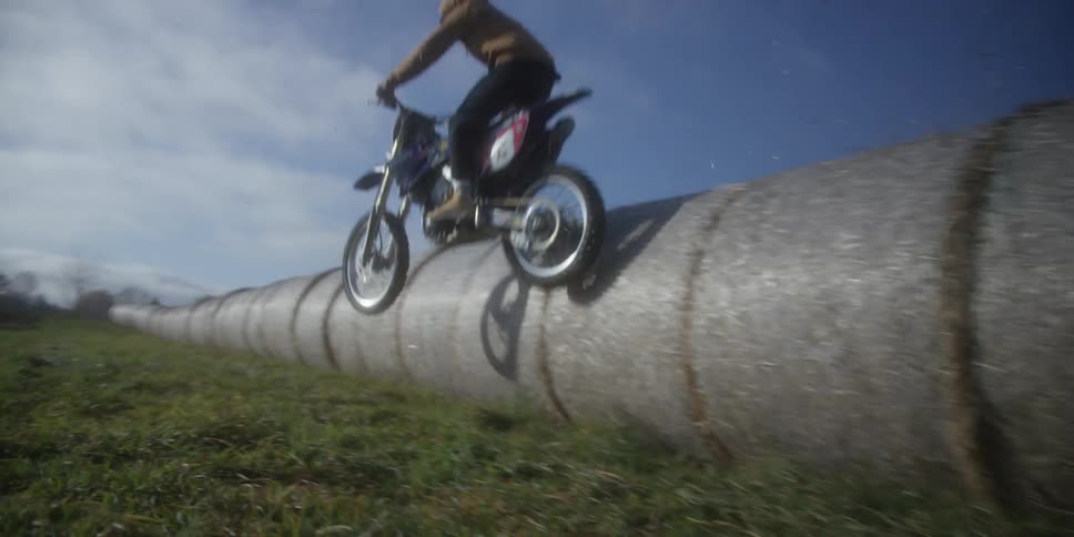 Motocross rider jumps over cylindrical pipe on grassy field beneath partly clear sky