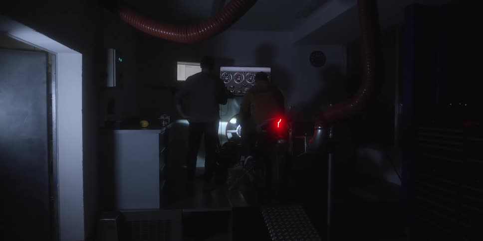 This is a short dark video showing what appears to be a motorcycle or vehicle being tested on a dynamometer in a dimly lit garage or shop two people are standing near the vehicle observing a screen displaying gauges or data with a red tail light visib
