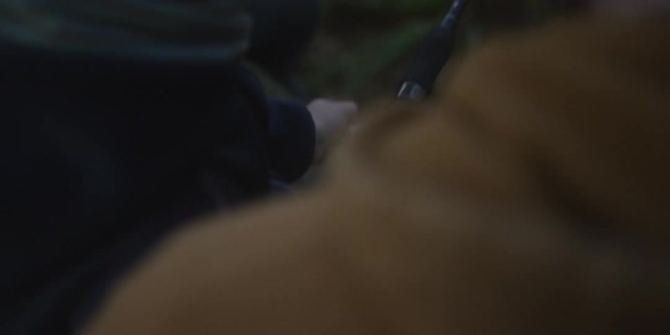 This is a closeup dimly lit view of a person operating a fishing reel on a rod turning the handle to wind in the line the scene is framed around the rod and reel with only parts of the angler and a dark outdoor background visible
