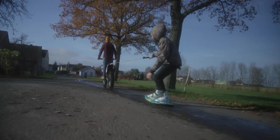A person is riding a dirt bike down a paved road pulling another person who is standing on a board possibly for wakeboarding or a similar activity on the wet road surfacethe scene takes place in a rural or suburban setting during autumn with a larg