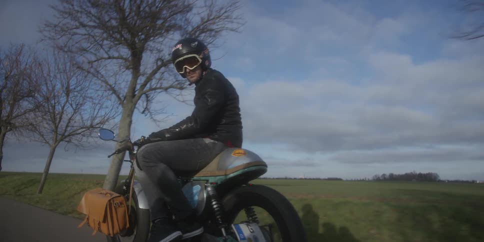 A motorcyclist riding a classic bike along a treelined rural road under a cloudy sky