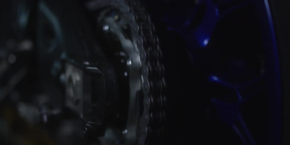 This is a closeup lowlight shot focusing on the drive chain of a motorcycle with a dark blue element visible to the right the chain and sprocket are stationary at first before the wheel begins to turn causing the chain to smoothly move