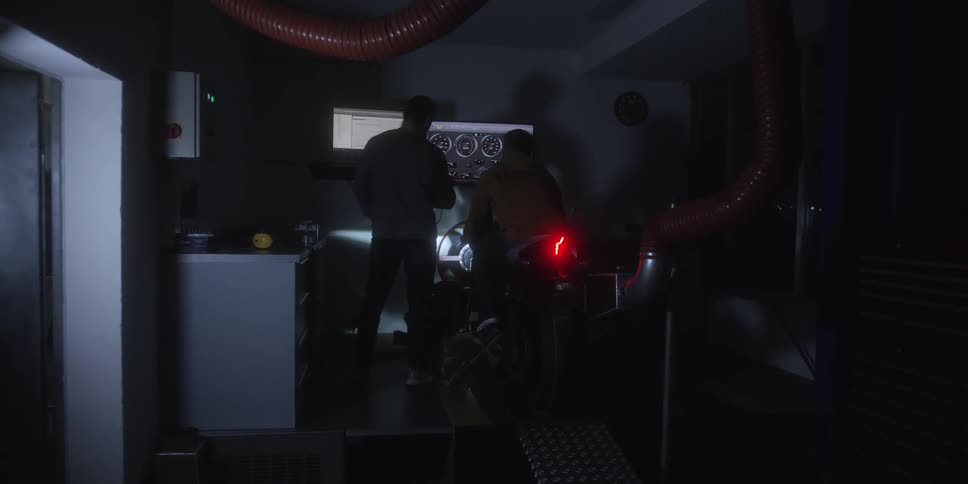 Two men work on a motorcycle with gauges on a screen in a dimly lit garage