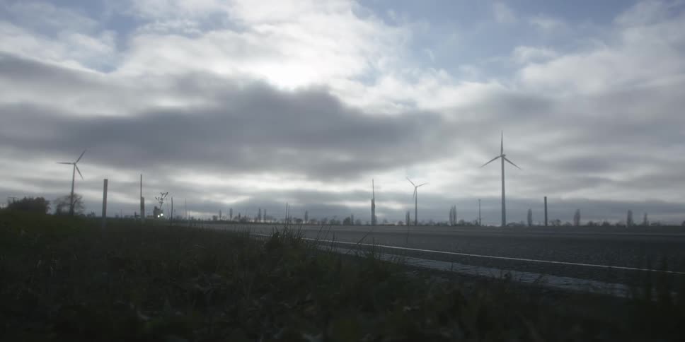 A motorcyclist rides along a rural road beneath a cloudy sky with wind turbines in the distance