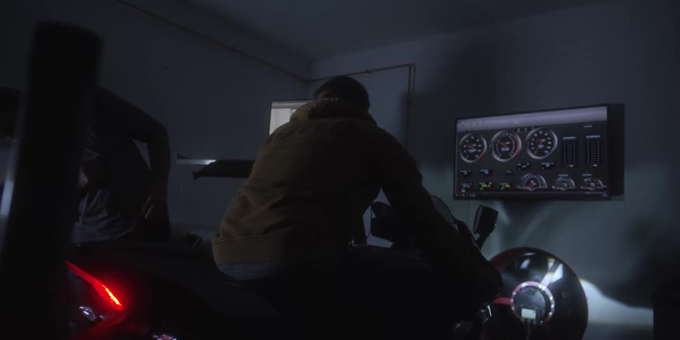 Two men test a motorcycle inside a dim garage while monitoring engine data on a screen