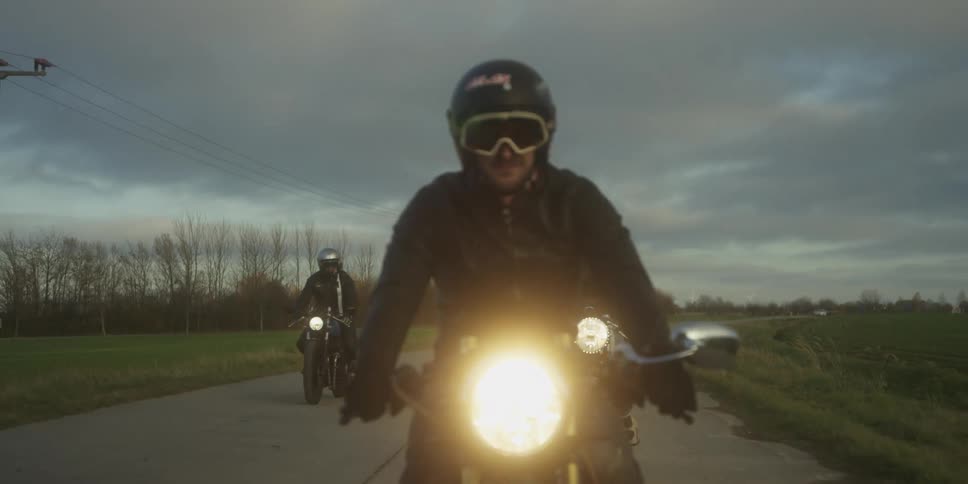 A group of motorcyclists rides together down a country road under a moody sky their bright headlights piercing the dusk as they approach in formation