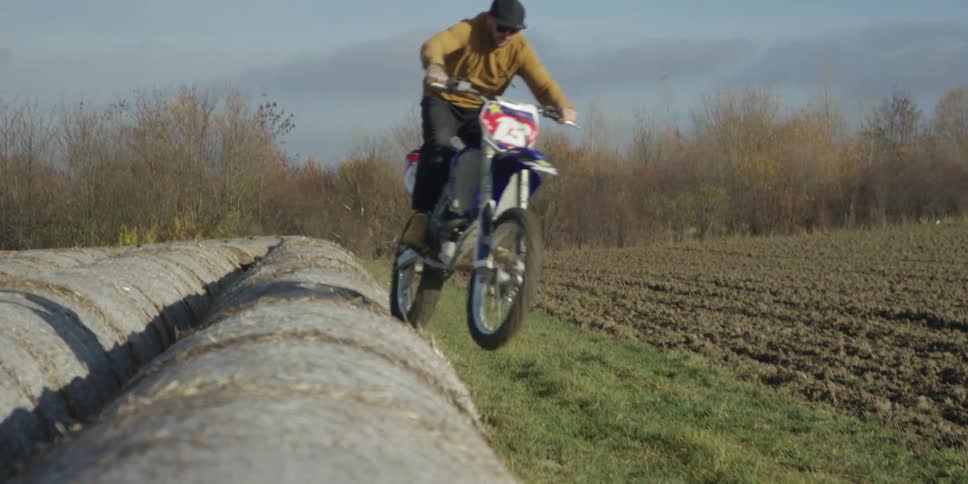A person rides a dirt bike across a row of large hay bales in an open rural field