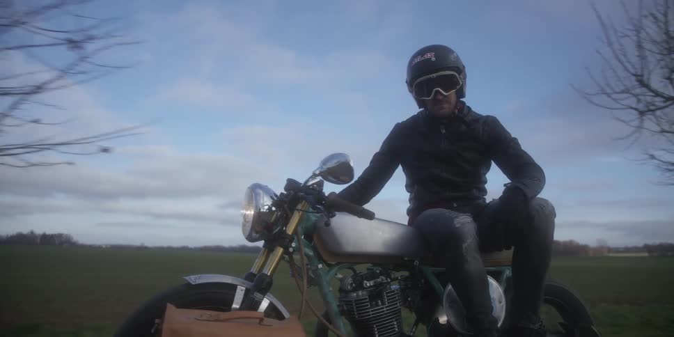 A motorcyclist wearing a helmet and goggles rides a motorcycle down a rural road past green fields and bare trees captured in a lowangle shot that highlights the bikes motion against the open sky