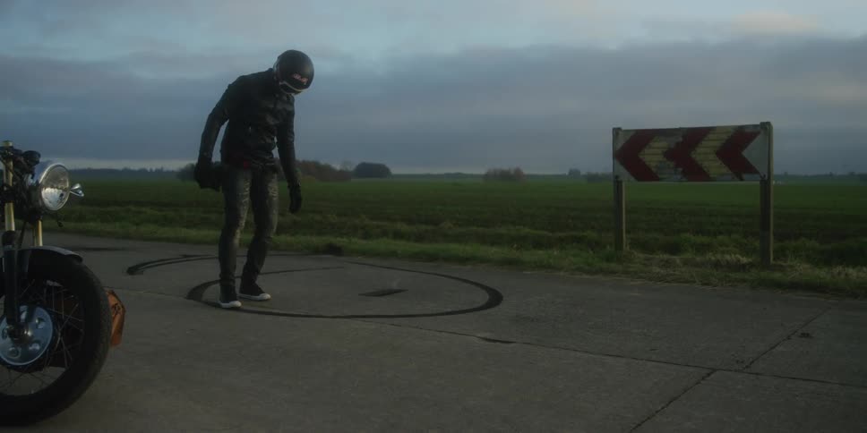 This video shows a person wearing a black motorcycle helmet and matching leather gear standing on a patch of pavement with skid marks possibly for a burnout a large chevron road sign indicating a sharp curve is visible in the background set against a c