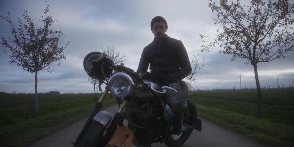 Man sits on his motorcycle parked on a rural road with a wind farm in the distance