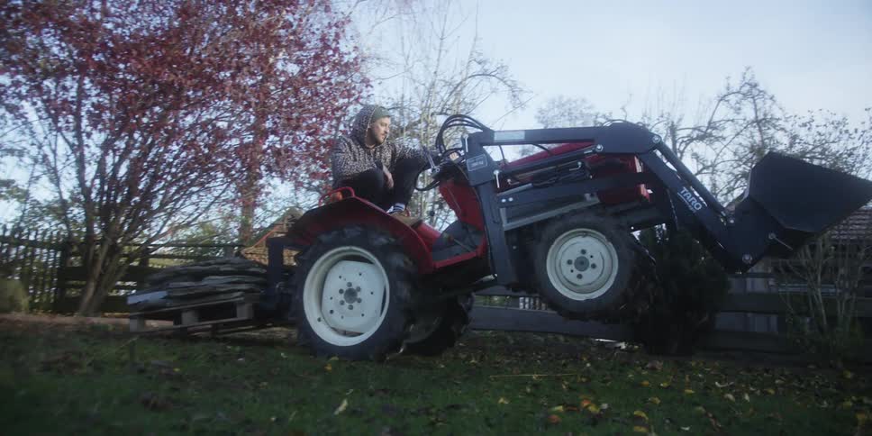 A person is operating a small red tractor with a front loader attachment in a yard attempting to lift or move a stack of wooden palletsthe front wheels of the tractor lift off the ground as the person engages the loader demonstrating a heavy lift be