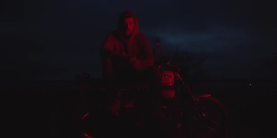This video features a man sitting on a motorcycle in a dark outdoor location illuminated primarily by a deep red light source he is wearing a hoodie and looking directly towards the camera which is slightly obscured by the lowlight conditions