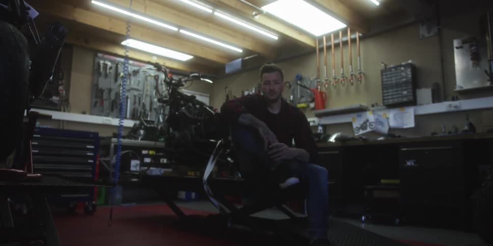 A man sits on a motorcycle lift looking directly at the camera inside a dimly lit workshop