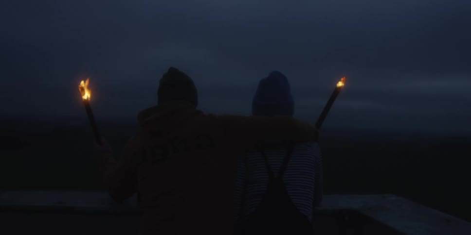 In the deep twilight two people holding flaming torches embrace and then triumphantly raise them high against the dark sky