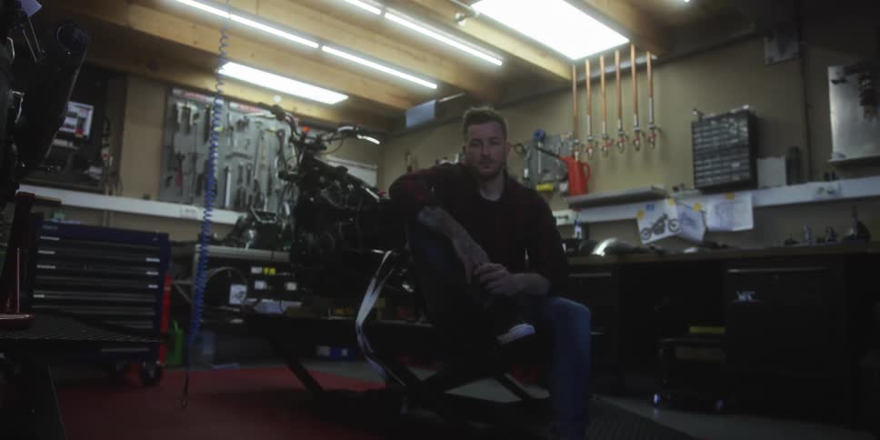 A mechanic sits on a motorcycle lift inside a brightly lit workshop