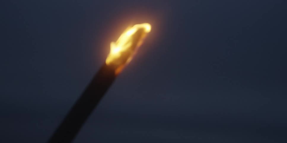 The video shows a closeup angled shot of a lit torch or flaming staff against a dark featureless background the orange and yellow flame flickers and glows brightly against the deep blue and gray of the night sky