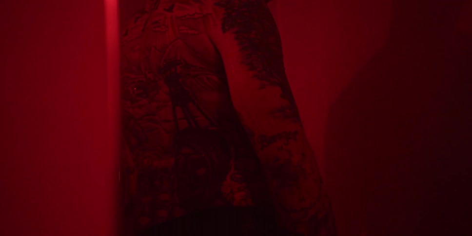 A man with tattoos over his body takes off his shirt inside a dark room facing the wall as red light illuminates the space