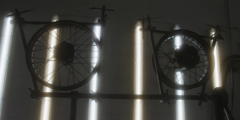 A slow shot pushes in on two wheels mounted on a metal rack silhouetted against glowing vertical fluorescent lights in a dark workshop