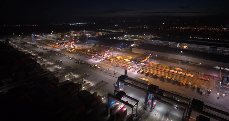 A wide high angle aerial view shows a brightly lit cargo depot with rows of shipping containers and trains at night