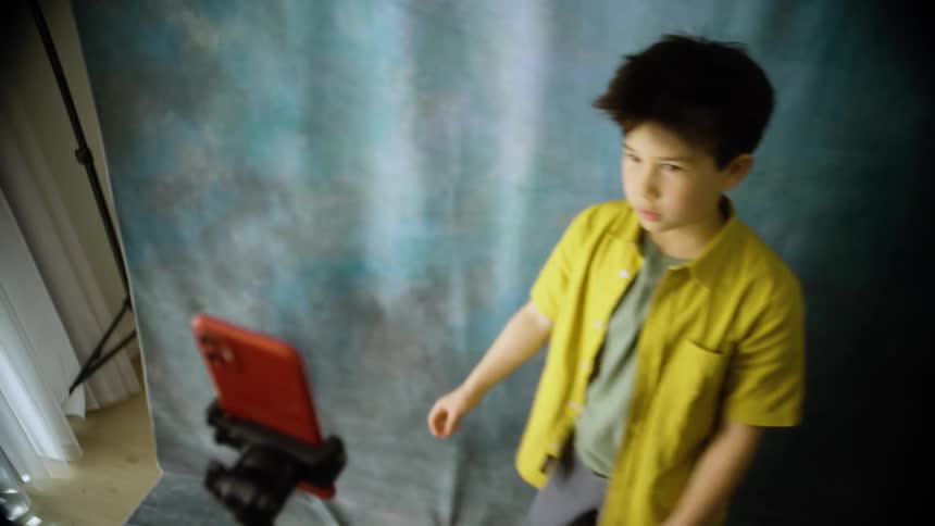A young boy in a yellow shirt stands in front of a blue backdrop performing expressive movements for a red smartphone mounted on a tripod