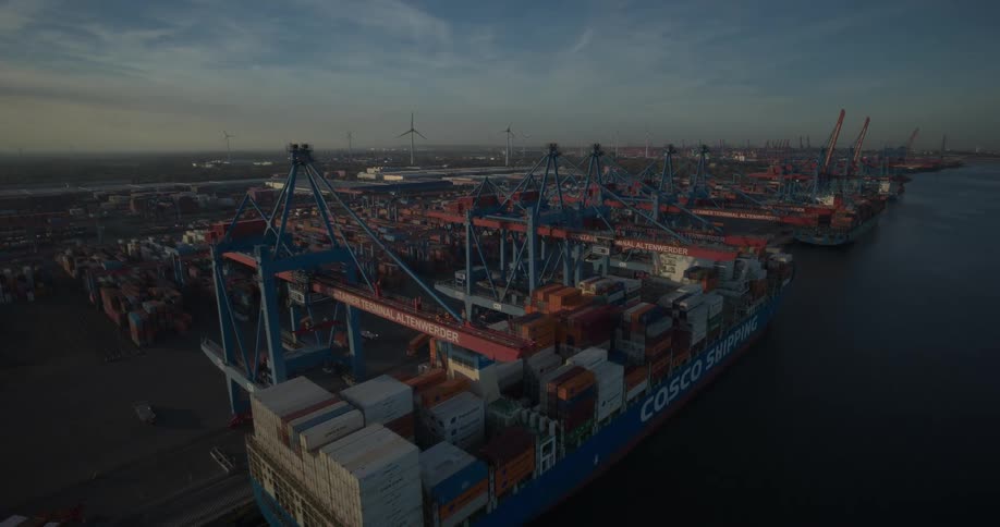 Ships at a loading dock with cranes on the coast and containers stacked on the ships and on shore as wind turbines spin in the distance