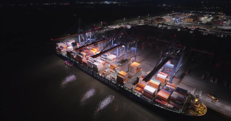 Cargo ship full of containers at a harbor with a metropolitan area on the background outdoors during dusk or dawn