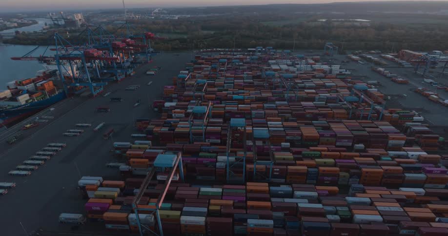 Containers stacked beneath blue cranes in industrial port at sunset with turbines in the background