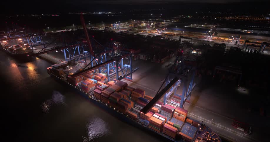 A high angle aerial view shows a massive container ship docked at a port at night actively being unloaded by large brightly lit port cranes over dark water
