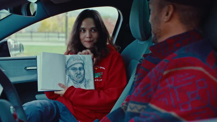 A woman in a red hoodie sits in a car and shows the man in the drivers seat a detailed pencil sketch of his face that she has drawn in her open notebook