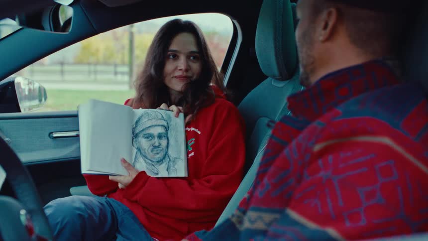 Teenager girl drawing on a notebook and showing to a man sitting next to her on a car while both laugh indoors during day time