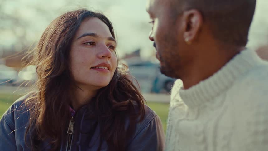 Young adult man wearing a white sweater sitting next to a young adult woman in a park and caressing her face while they talk outdoors during day time