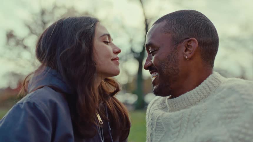 Young adult woman kissing a young adult man in the forehead while both smile sitting on the ground at a park on a suburban area outdoors during day time