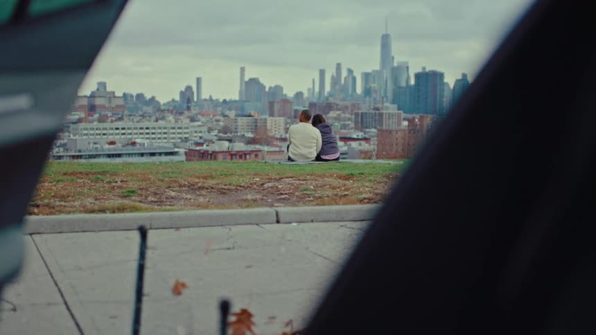 A couple sits closely together on a grassy hill overlooking a vast urban city skyline beneath a cloudy sky framed by dark foreground objects