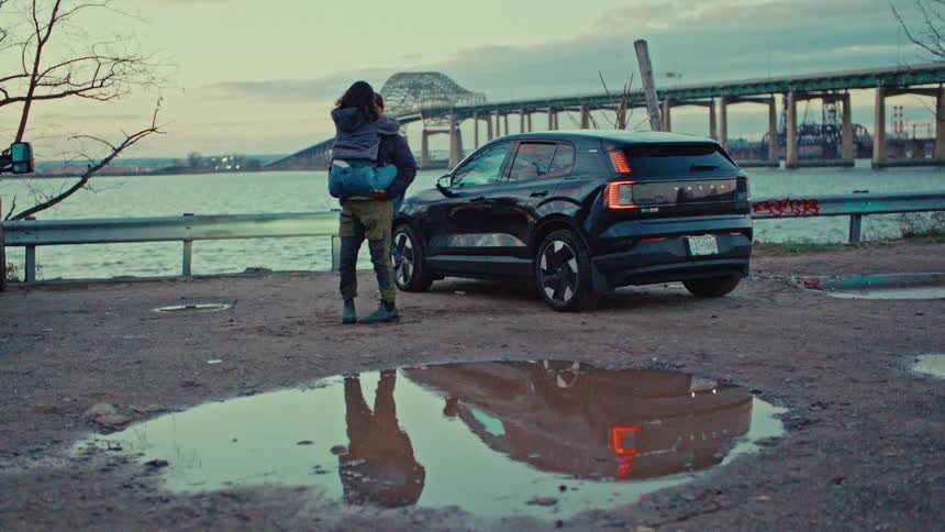 A young man stands by his car as a young woman runs to him they hug happily beside the car and he lifts her up with a bridge over a river in the background at sunset