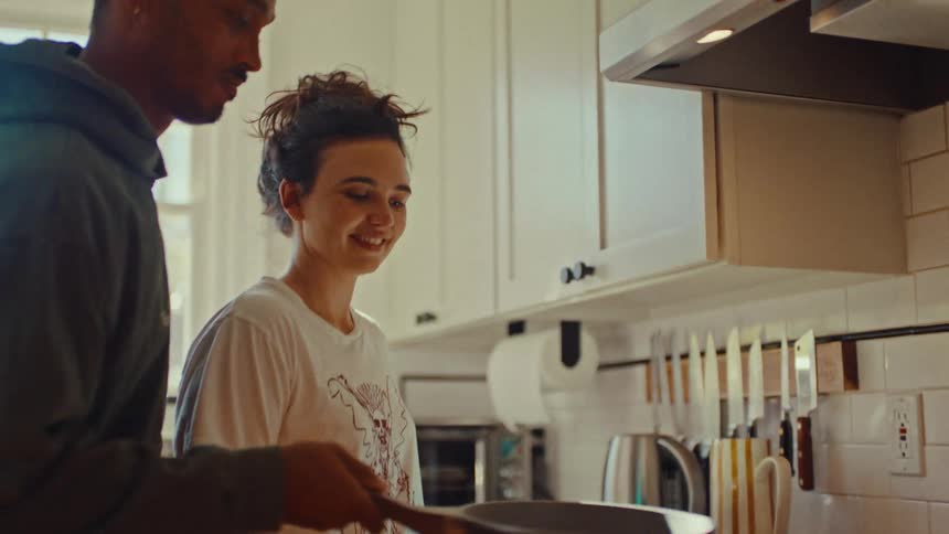Young adult woman watchng and laughing while a young adult man flips a pancake on top of the stove in a kitchen indoors during day time