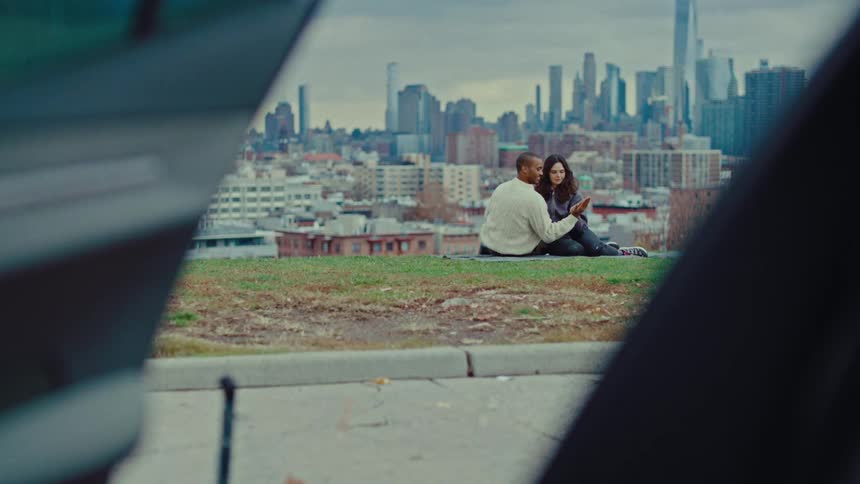 From inside a car a man and woman are seen holdings hands in a park near a metropolis