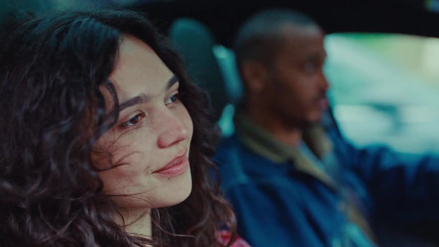 A man and a woman in the front seat of a car exchange a friendly glance and smile while driving during the day