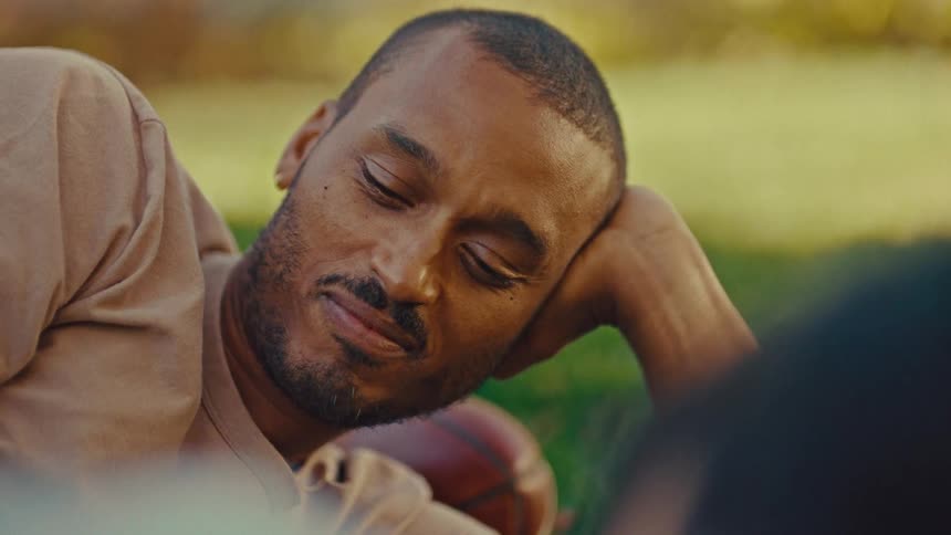A man wearing a light brown shirt lies smiling on a grassy lawn resting his head on his hand