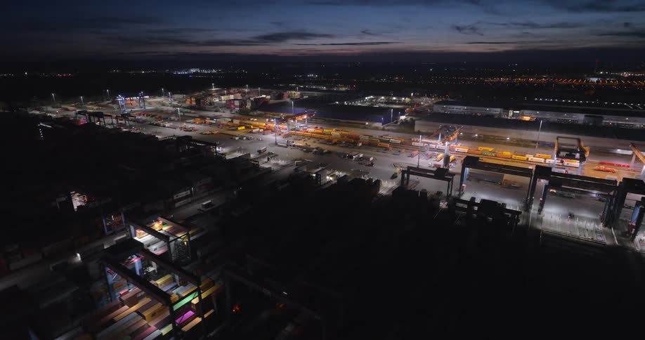 An aerial night view glides over a massive intermodal logistics hub revealing brightly lit rows of stacked cargo containers railway tracks and towering cranes working efficiently under the dark sky