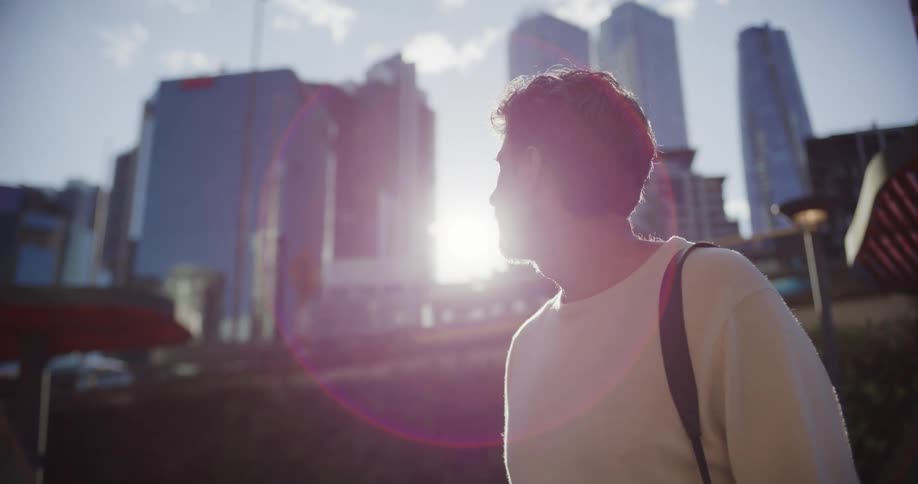 This is a lowangle shot of a man standing outdoors in a city looking up and away from the camera the bright sunlight creates a strong lens flare backlighting him against a backdrop of modern skyscrapers