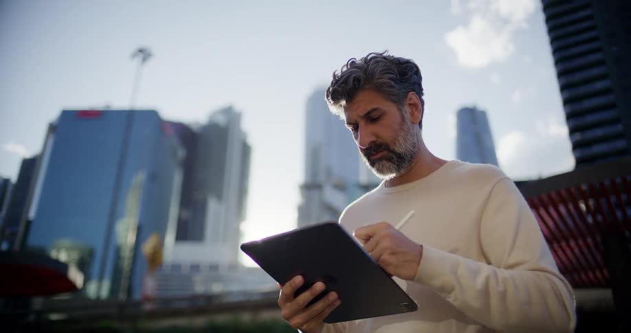 Man stands outside downtown working on a tablet computer with towers and skyscrapers in the distance