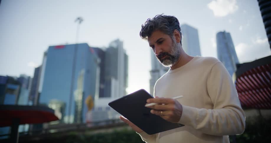 Man stands outside downtown working on a tablet computer with towers and skyscrapers in the distance