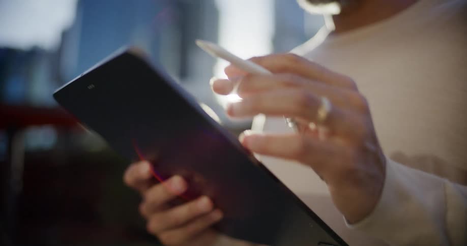 The video is a closeup shot of a persons hands holding a dark tablet or device and using a white stylus to interact with the screen the person is wearing a lightcolored longsleeved shirt and a ring with bright sun flare dominating the background