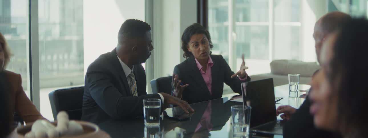 Small group of people from different ages and ethnicities sitting around a glass table on an office talking at a business meeting indoors during day time