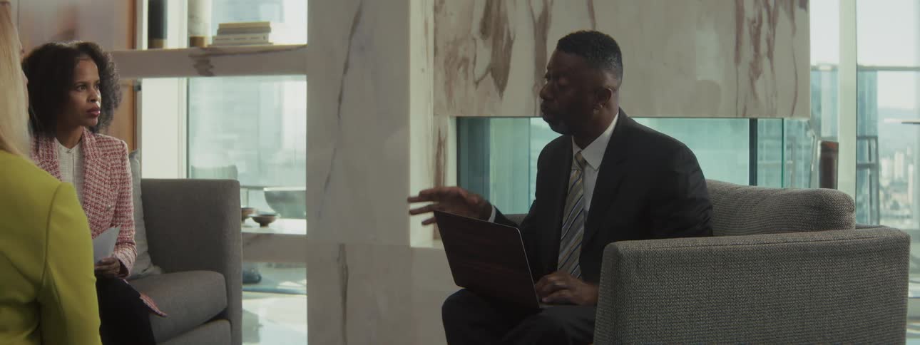 A professional man in a suit is sitting on a couch or chair talking and gesturing with his hand while holding a laptophe appears to be in a business meeting or consultation with a woman who is seated across from him holding papers