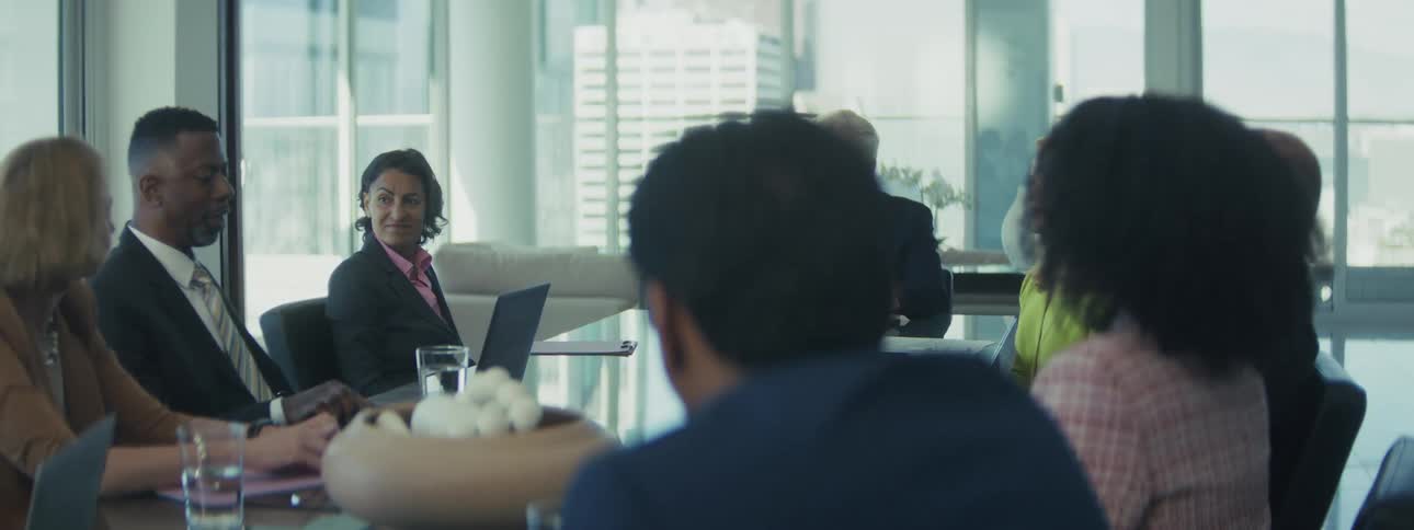 An overtheshoulder shot captures a diverse group of business professionals sitting around a large glass conference table in a modern sunlit office with a blurred cityscape view engaged in a conversation