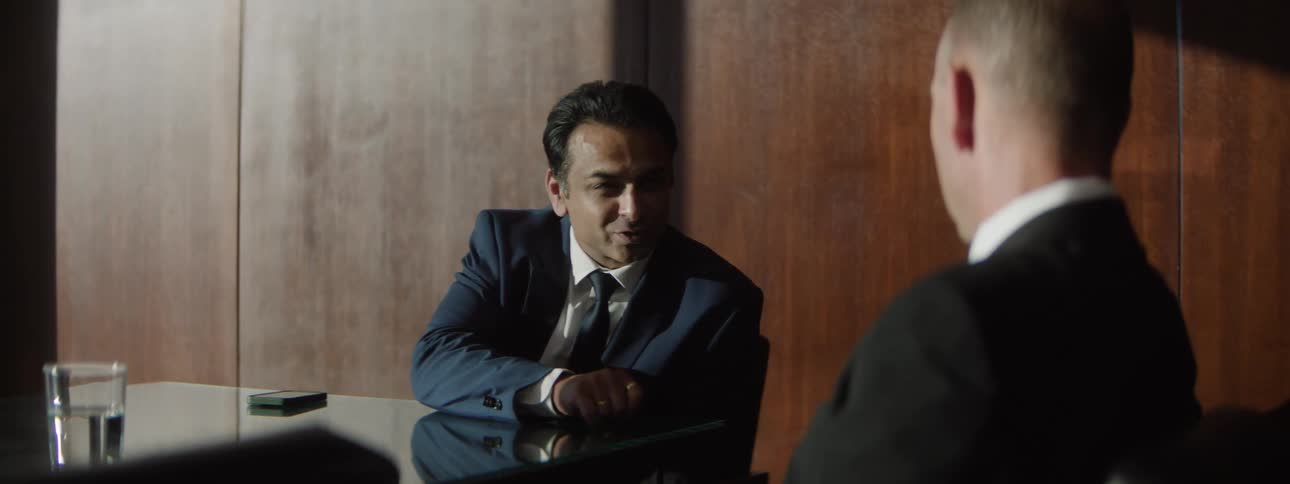 Two are sitting at a table in an office setting talking intently then they stand up a cityscape is in view outside the window