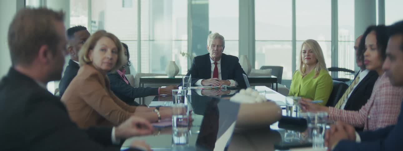A diverse group of professionals engage in a boardroom meeting around a large table with a city skyline backdrop